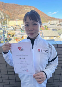 Woman on a balcony holding a certificate toward the camera with a mountainous town in the background.