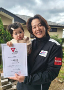 Woman in a black jacket smiles while holding a young child and a certificate outdoors in front of a house garden area.