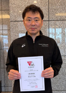 Asian man in a black zip-up jacket holding a certificate, standing in front of a tiled granite wall.