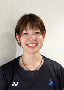 Smiling woman with short brown hair wearing a black athletic shirt with team logos, standing against a light wall.