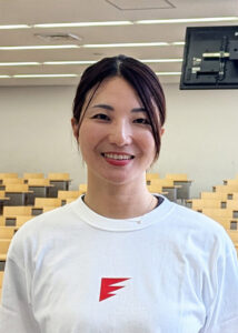 Smiling woman in a white t-shirt with a red lightning logo, standing in a classroom with wooden desks and a mounted screen above.